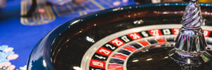 Vibrant casino table with roulette in motion, with casino chips, tokens, the hand of croupier, dollar money and a group of gambling rich people playing bet, blue poker table and deck of cards